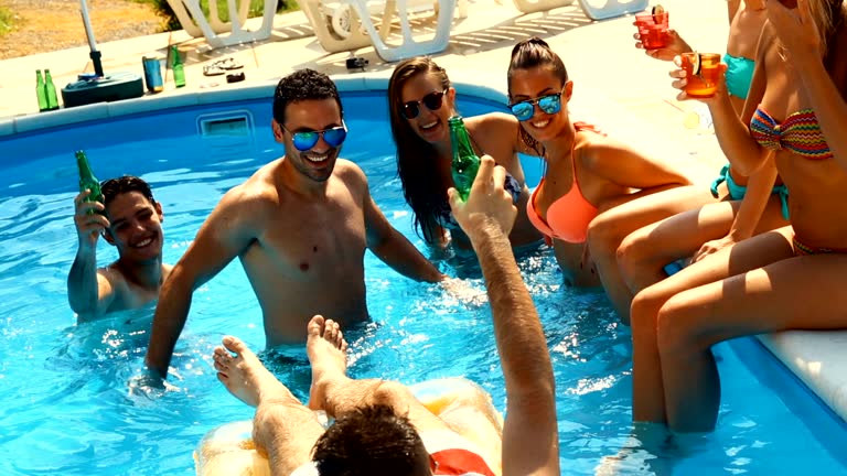 Closeup side view of four girls and three guys relaxing and enjoying in a swimming pool on a hot summer day. They are waist deep in the water, having cocktails and dancing. Stabilized dolly shot.